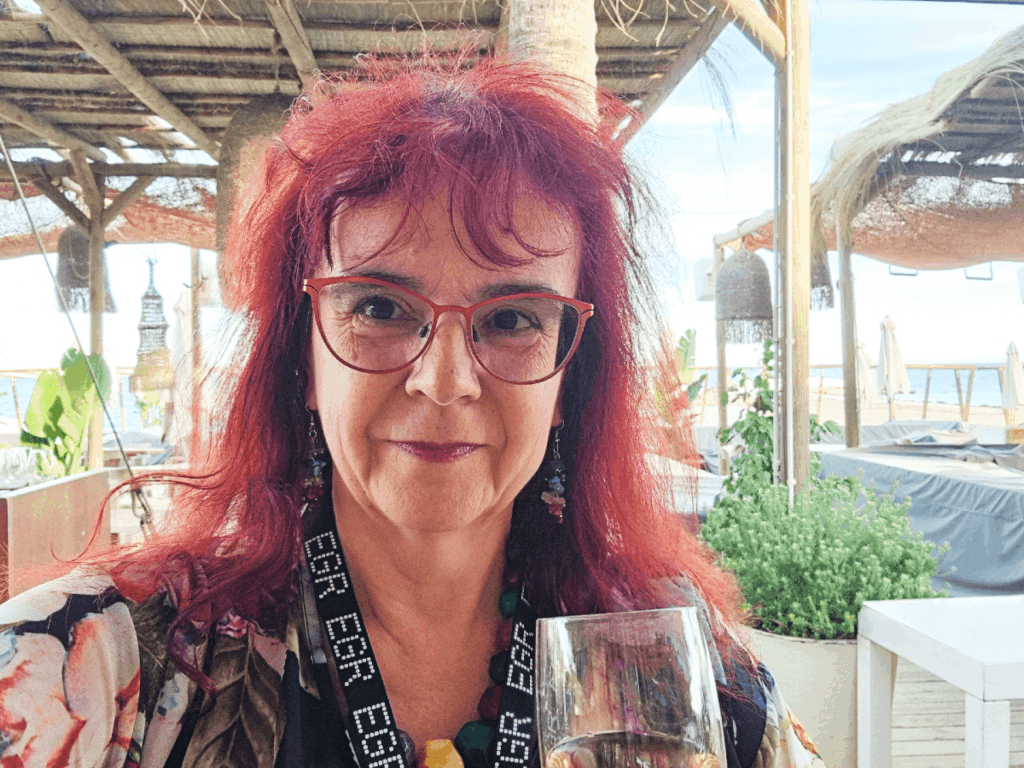 Sue Dawson stands in the Chiringuito beach bar at the Puente Romano Resort, Marbella. A middle aged lady with dark red hair, she is holding a champagne glass and smiling